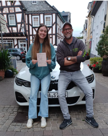 Fahrschülerin zeigt stolz ihre bestandene Prüfungsbescheinigung neben Fahrlehrer Norman Seibert